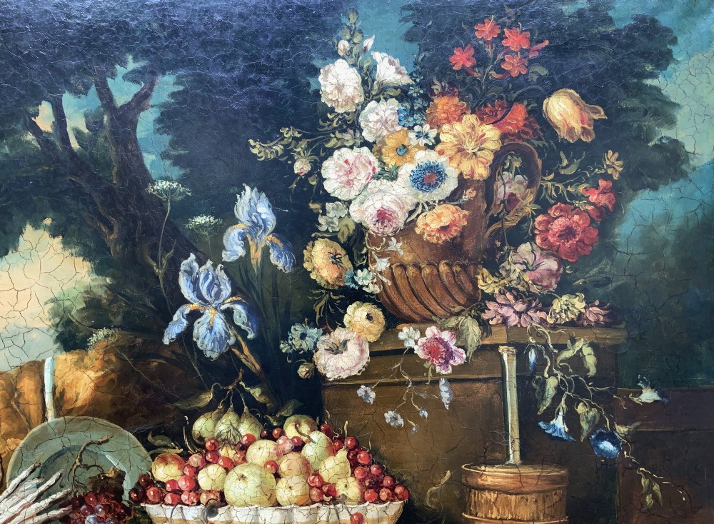 A classical oil painting displaying a bowl of fruit, a urn with flowers, asparagus, and melons set against a dark landscape backdrop within a carved silver-grey frame. View VII