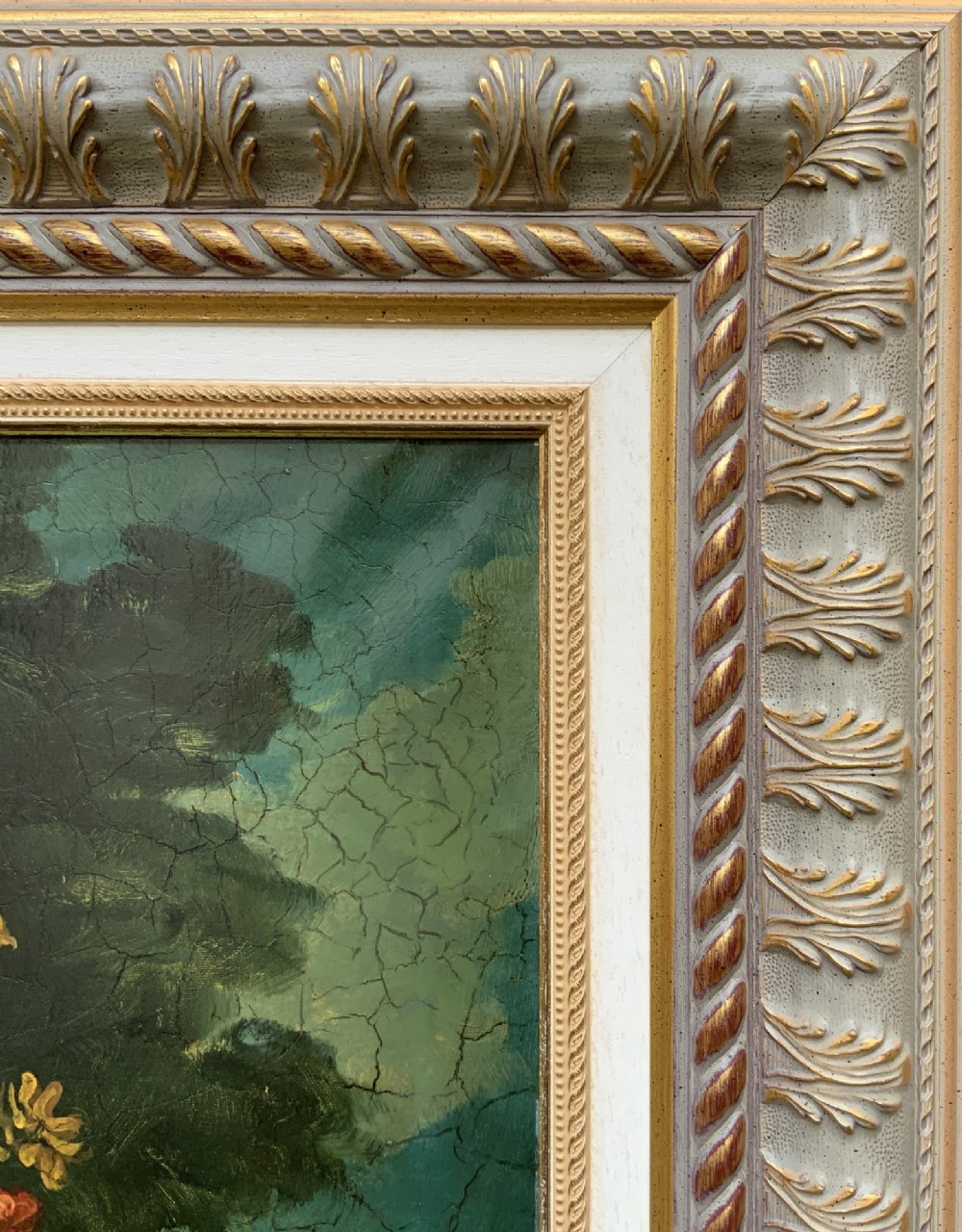 A classical oil painting displaying a bowl of fruit, a urn with flowers, asparagus, and melons set against a dark landscape backdrop within a carved silver-grey frame. View X