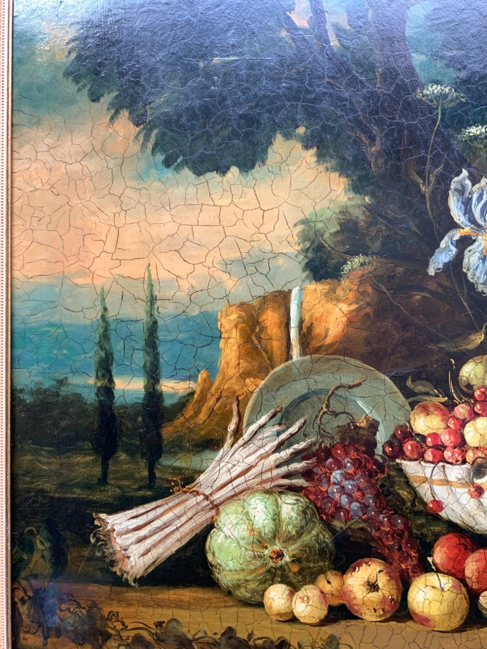 A classical oil painting displaying a bowl of fruit, a urn with flowers, asparagus, and melons set against a dark landscape backdrop within a carved silver-grey frame. View XIII