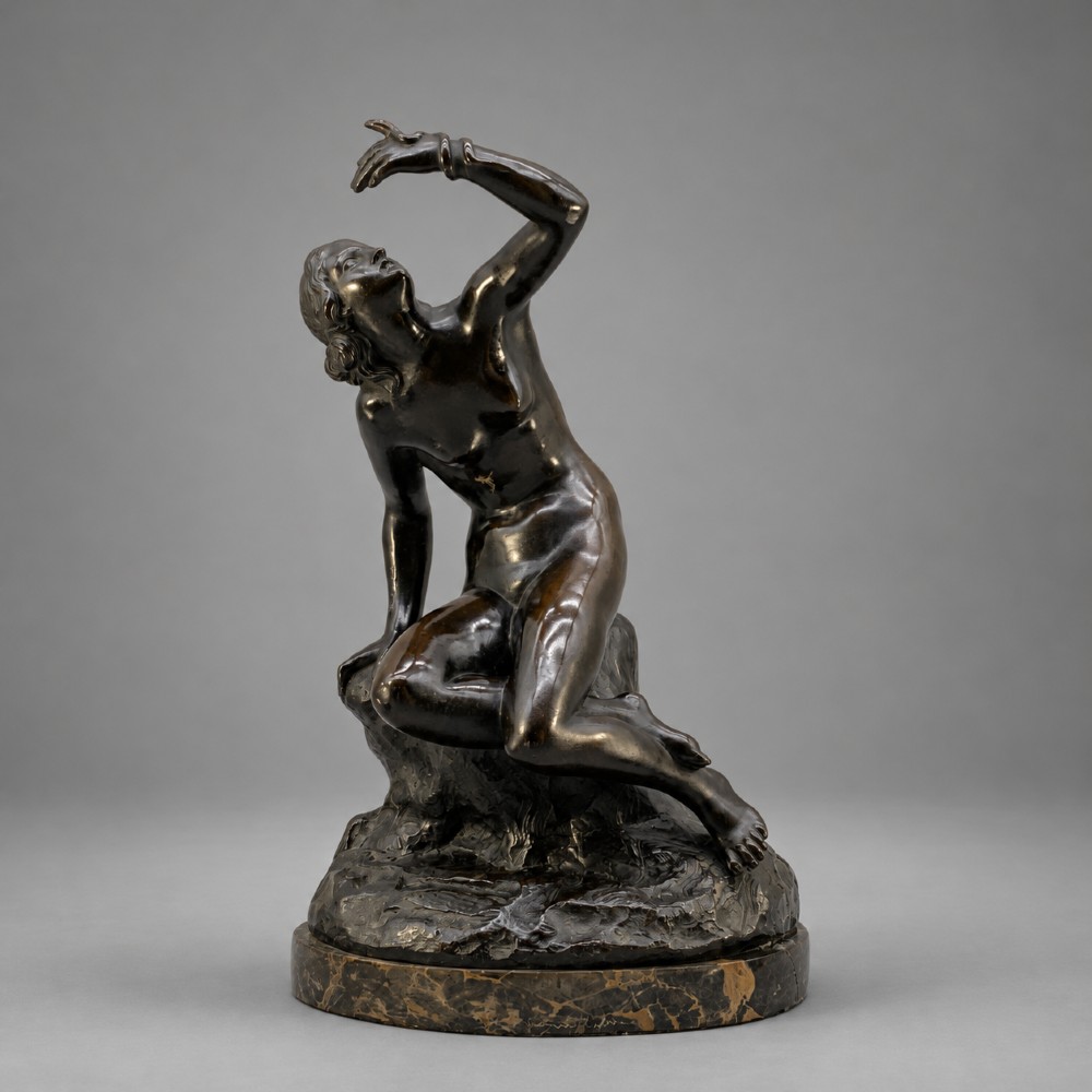 A patinated dark bronze sculpture depicting a classical female figure reaching upward while seated on a rocky outcrop atop a circular marble base.