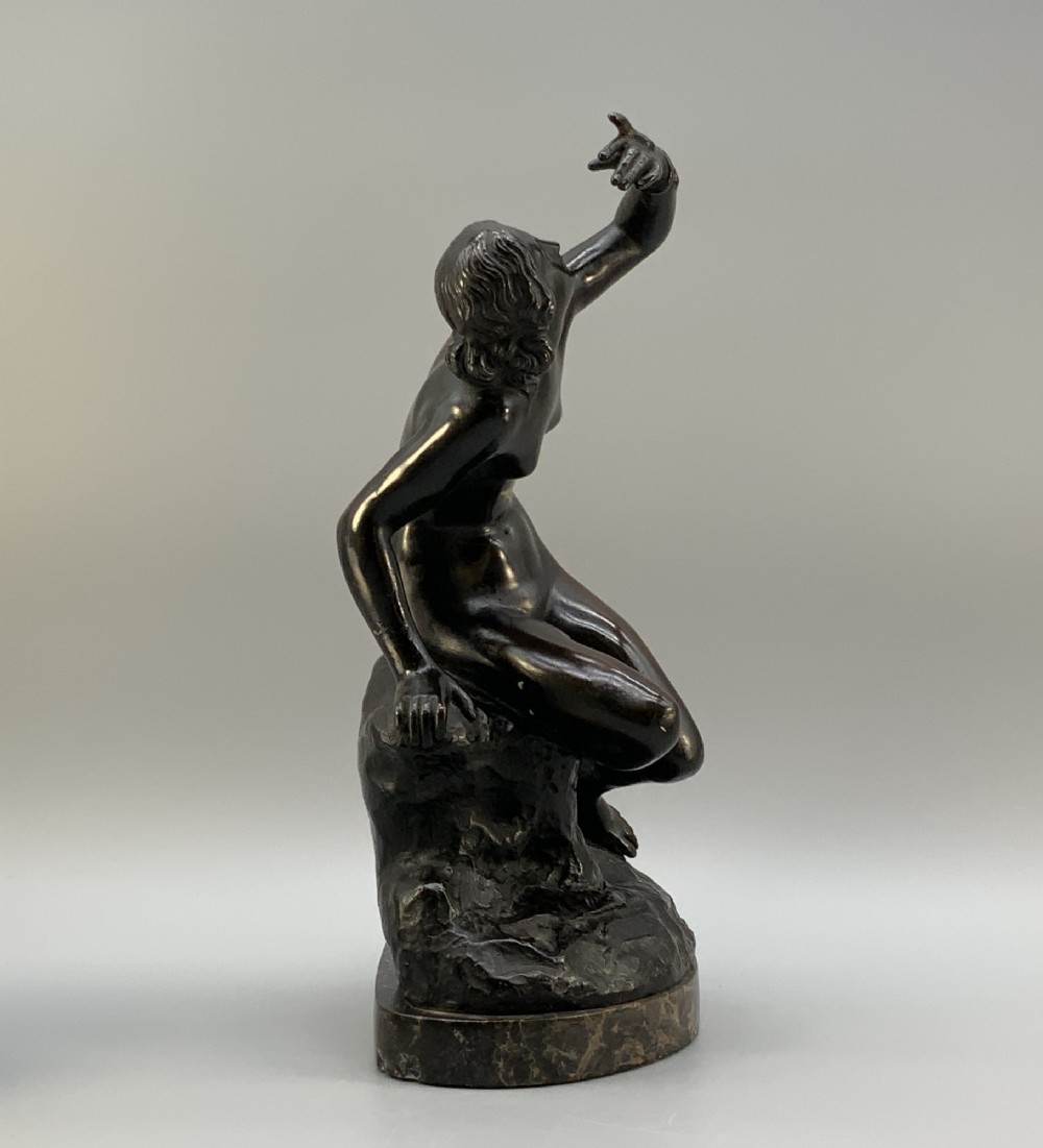 A patinated dark bronze sculpture depicting a classical female figure reaching upward while seated on a rocky outcrop atop a circular marble base. View V