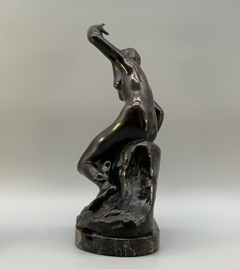 A patinated dark bronze sculpture depicting a classical female figure reaching upward while seated on a rocky outcrop atop a circular marble base. View VII