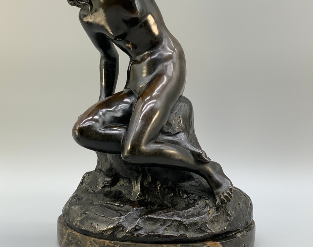 A patinated dark bronze sculpture depicting a classical female figure reaching upward while seated on a rocky outcrop atop a circular marble base. View X