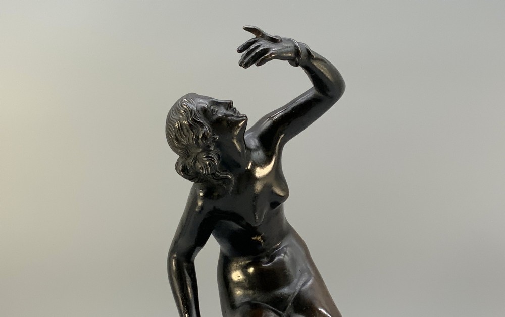 A patinated dark bronze sculpture depicting a classical female figure reaching upward while seated on a rocky outcrop atop a circular marble base. View XII