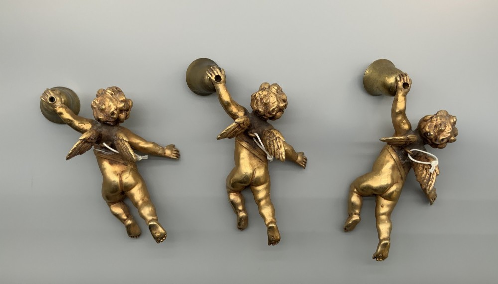 A set of three gilt metal wall sconces crafted in the shape of winged cherubs holding bell-shaped light sockets. View II