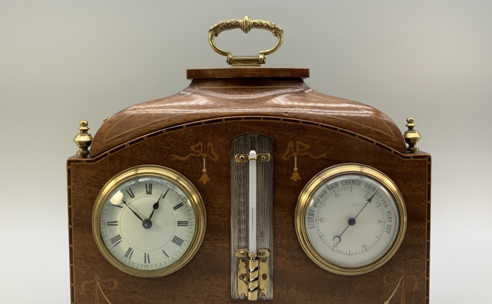 An Edwardian mahogany desk compendium featuring an inlaid floral swag motif, central thermometer, clock dial, barometer dial, and top brass handle. View V