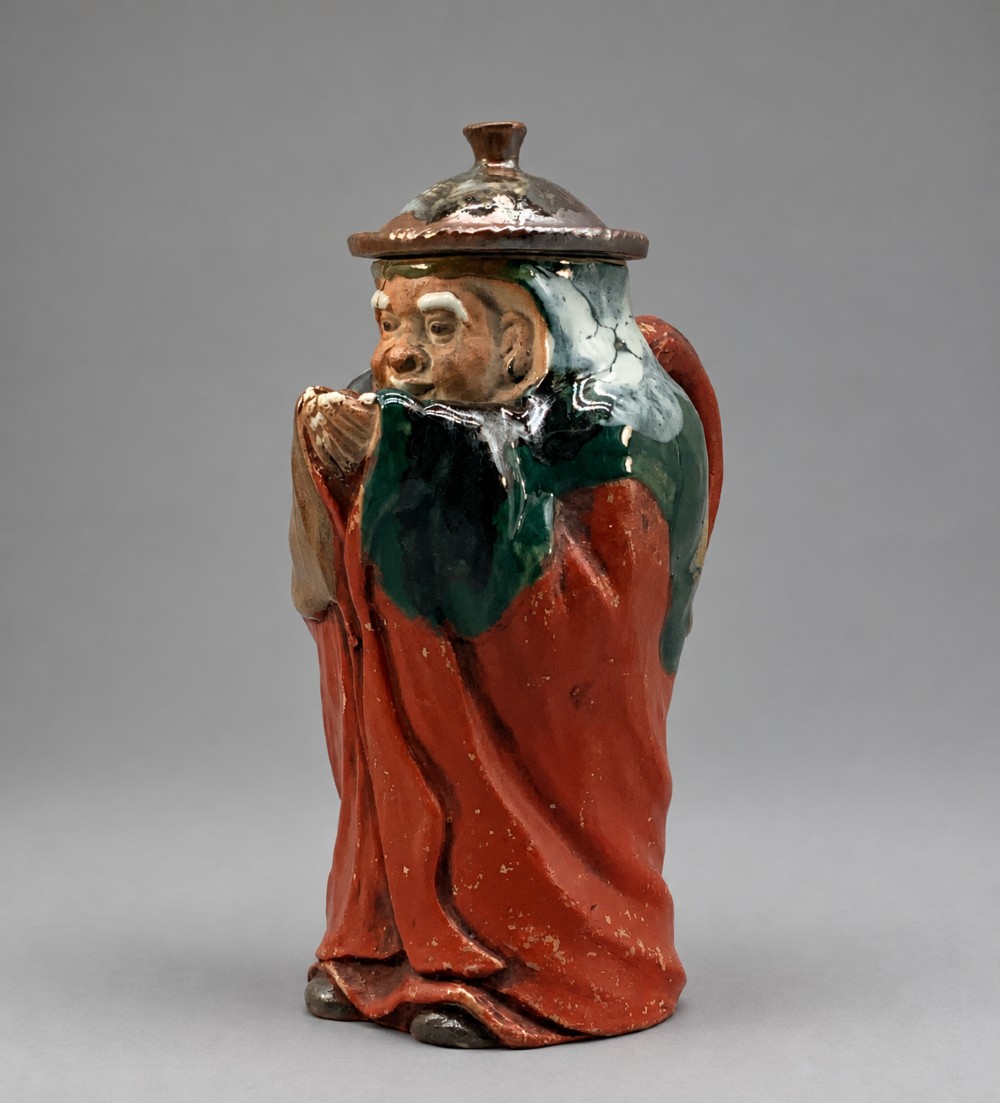 An antique Japanese Sumida Gawa lidded pottery jug sculpted in the form of a robed figure with red attire, green glaze drips, and a matching fitted lid.