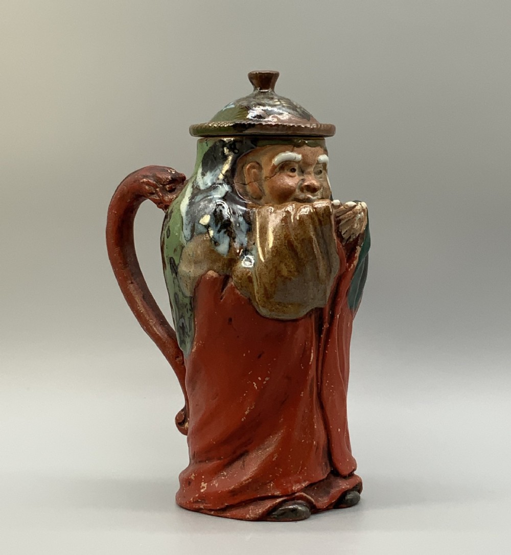 An antique Japanese Sumida Gawa lidded pottery jug sculpted in the form of a robed figure with red attire, green glaze drips, and a matching fitted lid. View V