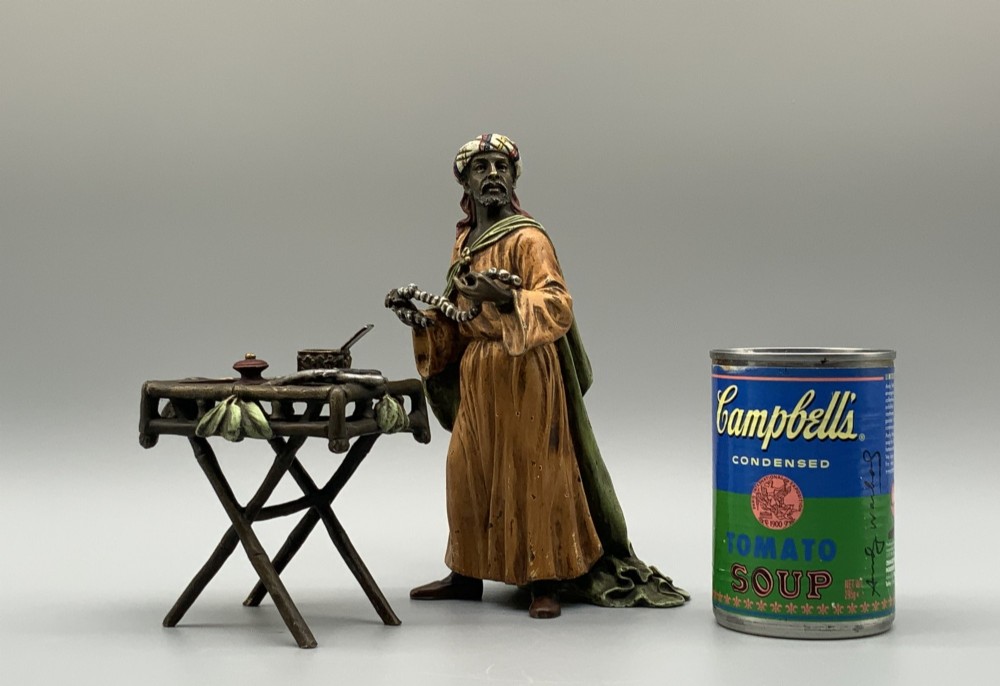 A cold-painted bronze Orientalist figure wearing a turban and ochre robe, standing next to a small table displaying wares. View II