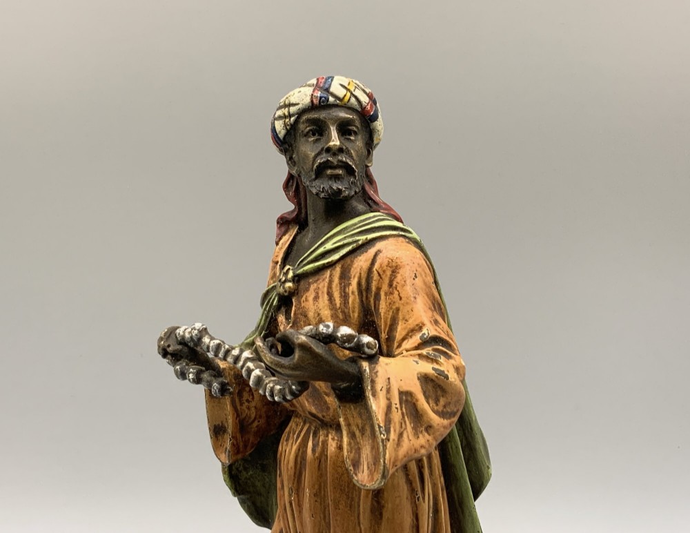 A cold-painted bronze Orientalist figure wearing a turban and ochre robe, standing next to a small table displaying wares. View III