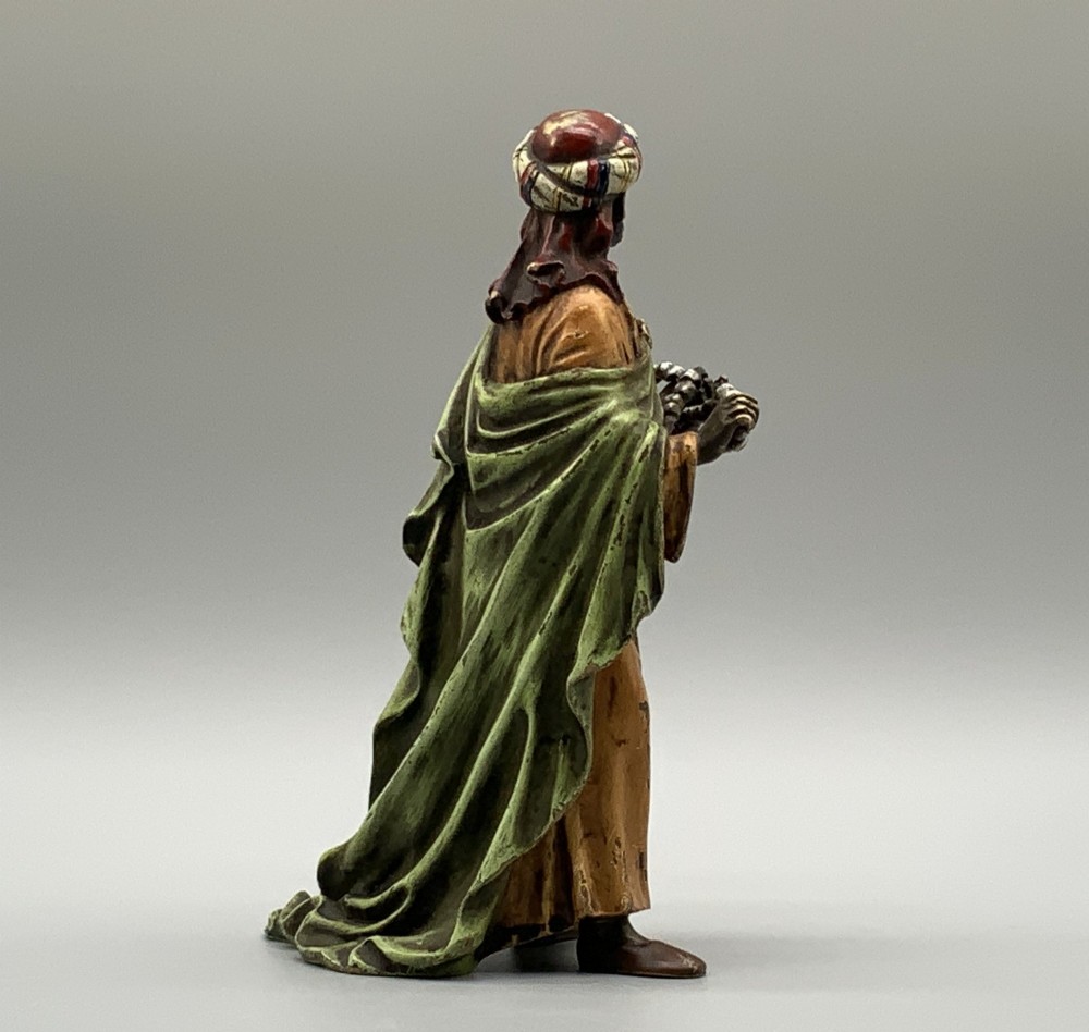 A cold-painted bronze Orientalist figure wearing a turban and ochre robe, standing next to a small table displaying wares. View VI