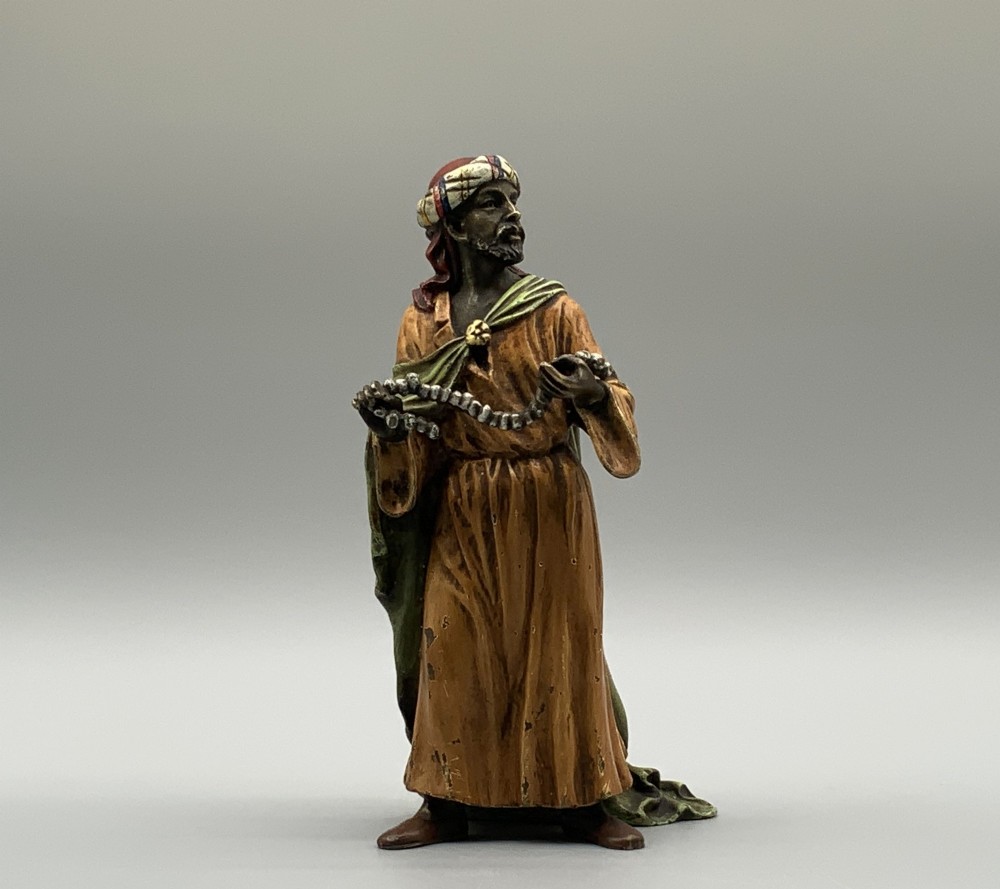 A cold-painted bronze Orientalist figure wearing a turban and ochre robe, standing next to a small table displaying wares. View VIII