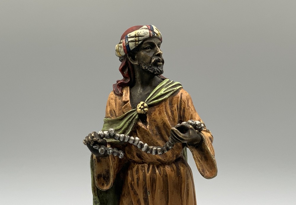 A cold-painted bronze Orientalist figure wearing a turban and ochre robe, standing next to a small table displaying wares. View IX