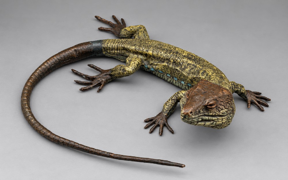 An antique cold-painted bronze sculpture depicting a realistic lizard with textured yellow and brown skin, blue accents, and a long curved tail.
