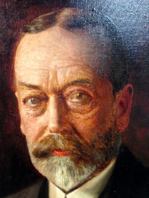 fig 2. A formal half-length oil portrait painting of King George V wearing a dark suit against a shaded brown background.