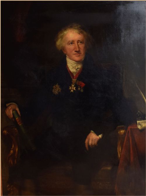 fig 5. An antique oil painting on canvas depicting a seated gentleman with white hair wearing a dark coat decorated with medals, holding a green book beside a red-draped table.