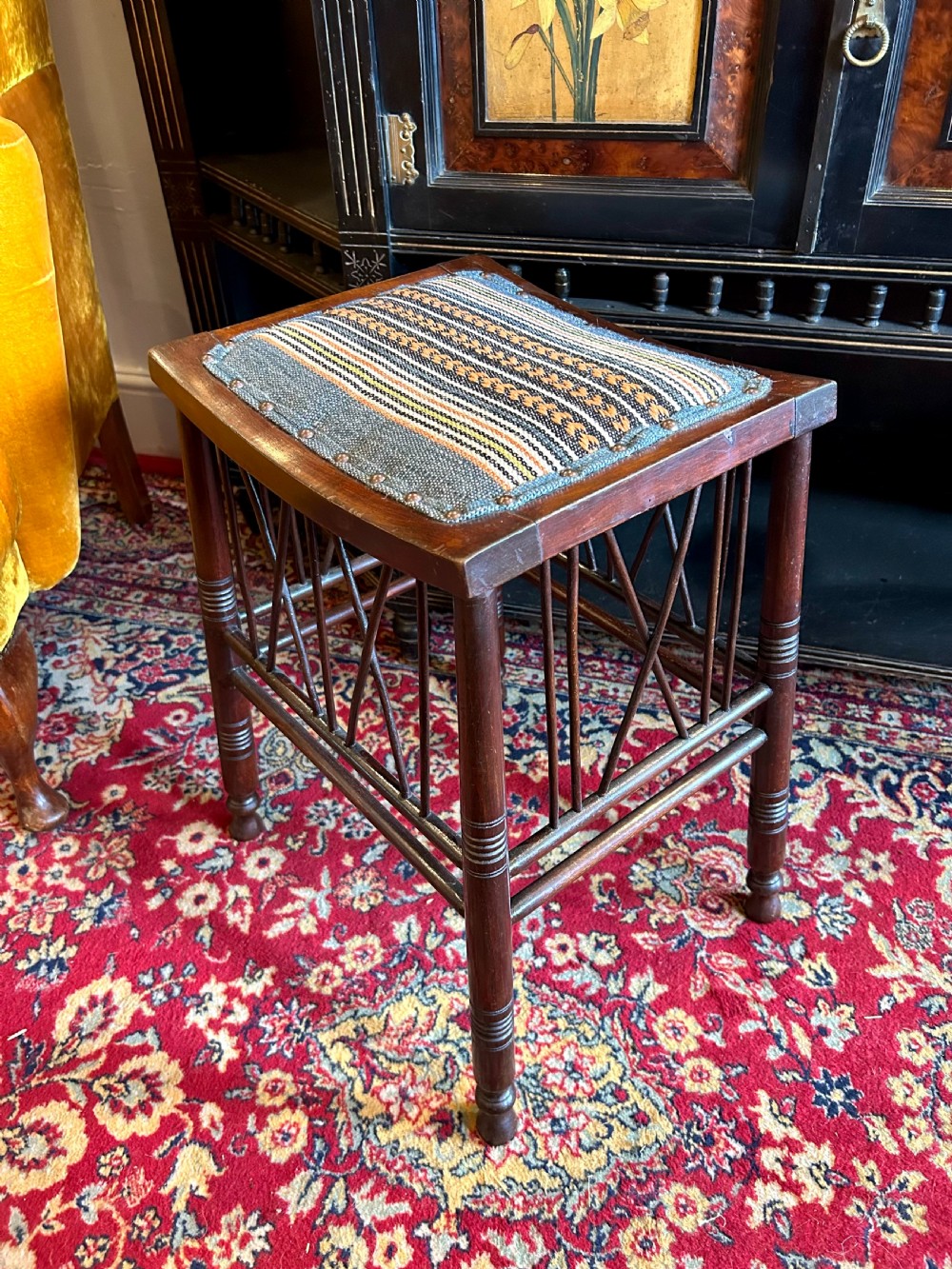 A rectangular mahogany Arts and Crafts Thebes style stool featuring turned legs, cross-braced spindle sides, and an upholstered woven patterned seat.