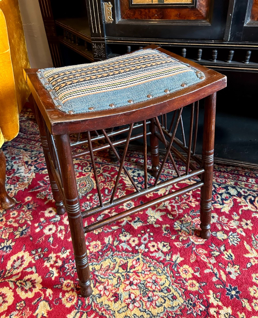 A rectangular mahogany Arts and Crafts Thebes style stool featuring turned legs, cross-braced spindle sides, and an upholstered woven patterned seat. View II