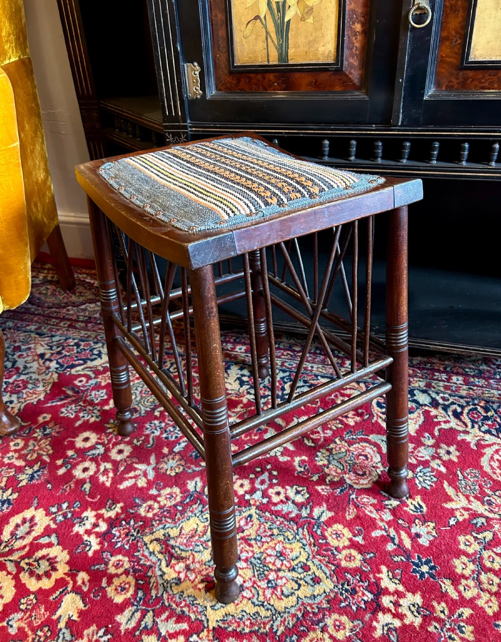 A rectangular mahogany Arts and Crafts Thebes style stool featuring turned legs, cross-braced spindle sides, and an upholstered woven patterned seat. View III