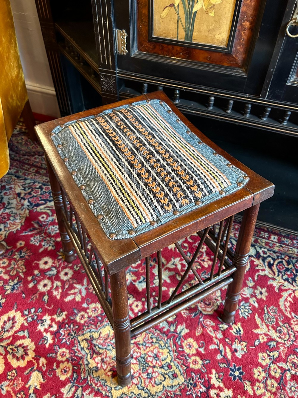 A rectangular mahogany Arts and Crafts Thebes style stool featuring turned legs, cross-braced spindle sides, and an upholstered woven patterned seat. View IV