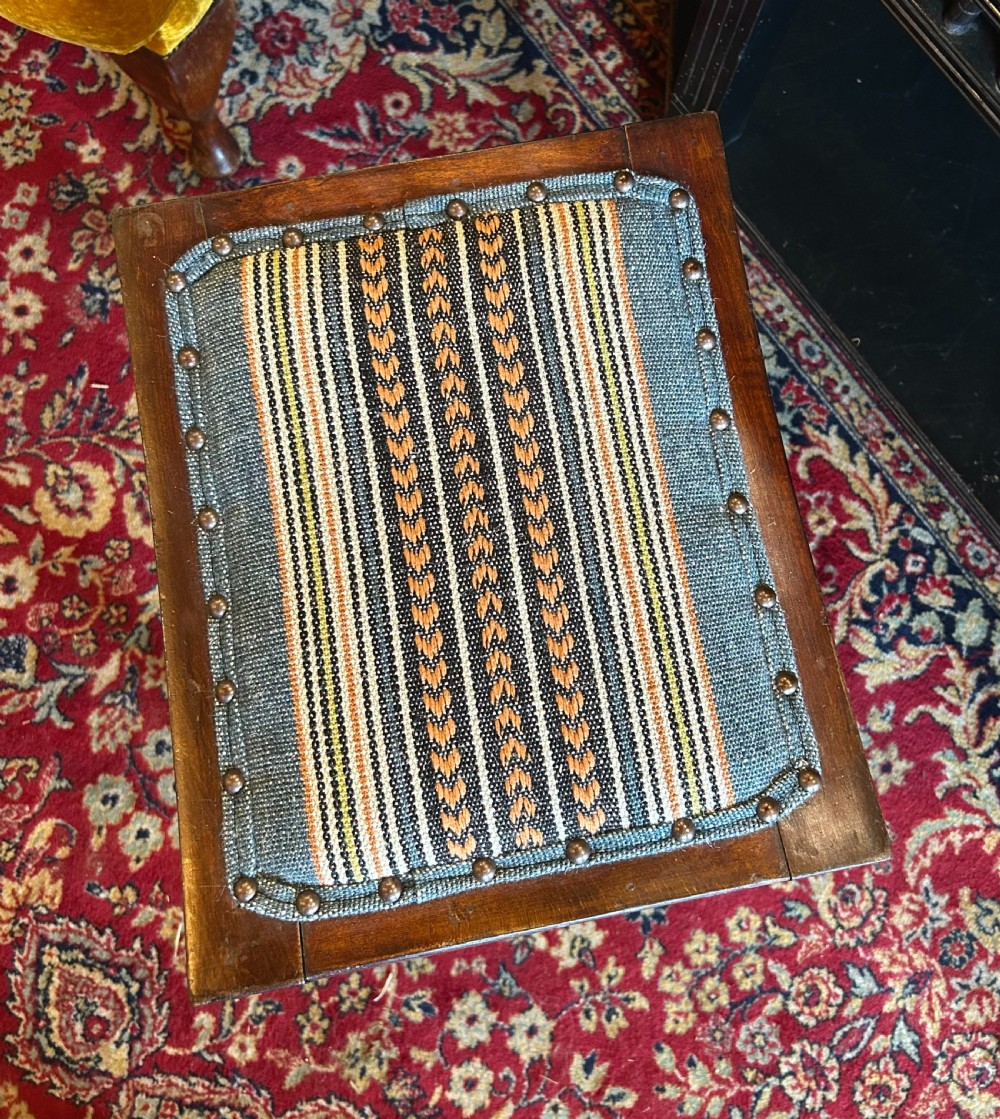 A rectangular mahogany Arts and Crafts Thebes style stool featuring turned legs, cross-braced spindle sides, and an upholstered woven patterned seat. View V