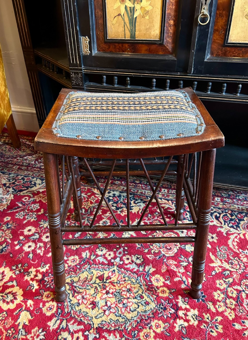 A rectangular mahogany Arts and Crafts Thebes style stool featuring turned legs, cross-braced spindle sides, and an upholstered woven patterned seat. View VI