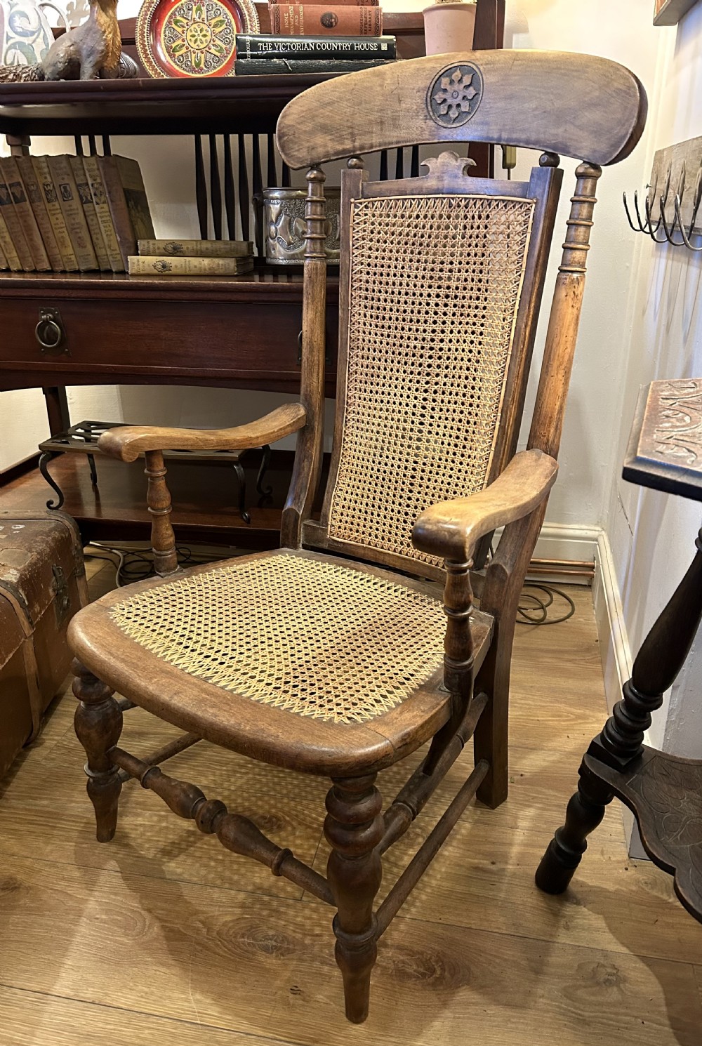 A rustic wooden armchair featuring a high back with a woven cane panel, a woven cane seat, curved armrests, and turned front legs with carved details on the top rail.