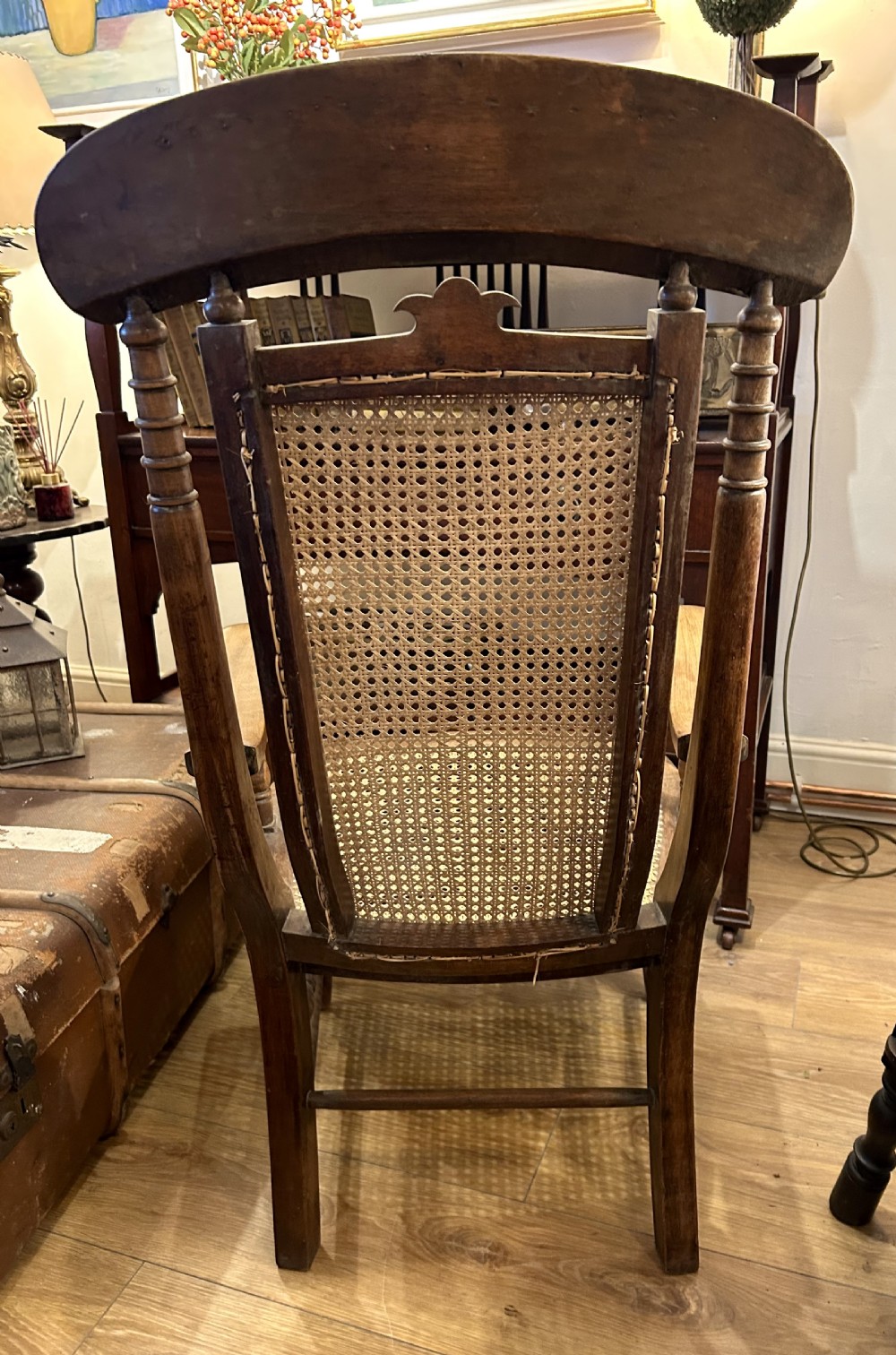 A rustic wooden armchair featuring a high back with a woven cane panel, a woven cane seat, curved armrests, and turned front legs with carved details on the top rail. View VI