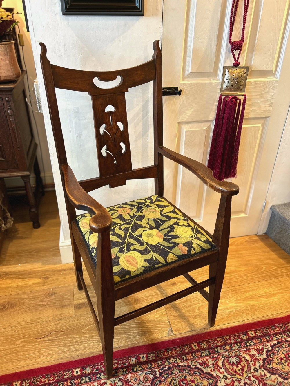 A dark oak Arts and Crafts armchair featuring a high back with a central floral inlaid splat, shaped armrests, and a botanical patterned upholstered seat. View II