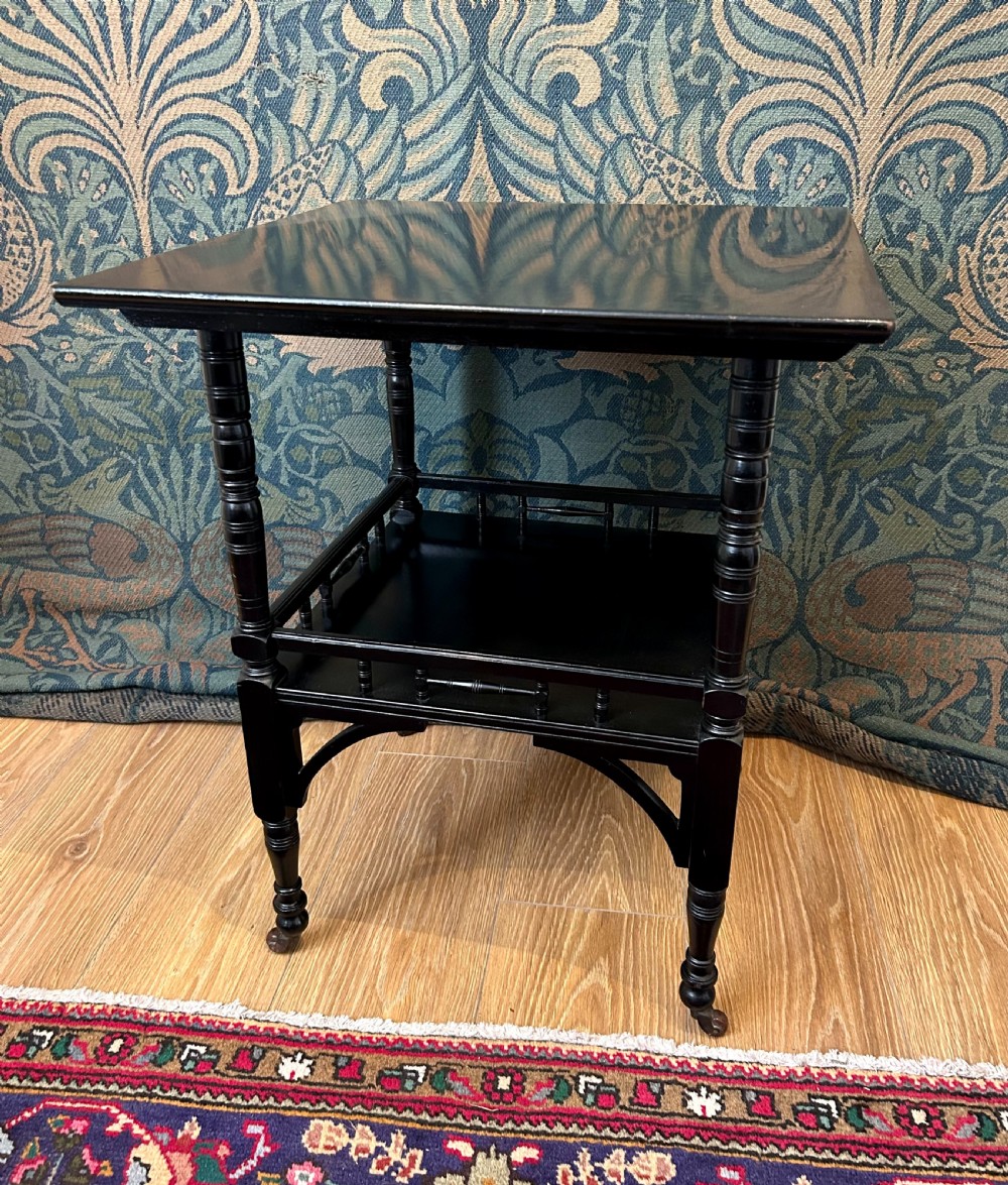 An Aesthetic Movement ebonised wooden side table featuring turned legs, a lower shelf with spindle railing, arched corner brackets, and brass castors.