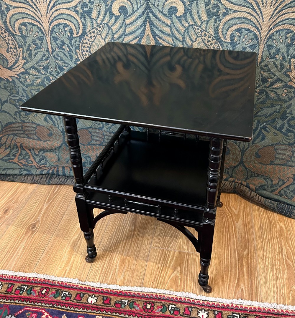 An Aesthetic Movement ebonised wooden side table featuring turned legs, a lower shelf with spindle railing, arched corner brackets, and brass castors. View II