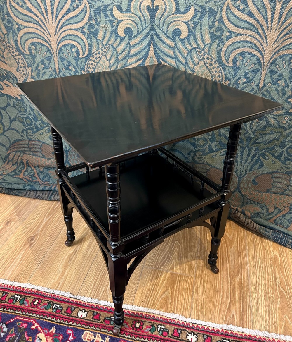 An Aesthetic Movement ebonised wooden side table featuring turned legs, a lower shelf with spindle railing, arched corner brackets, and brass castors. View III