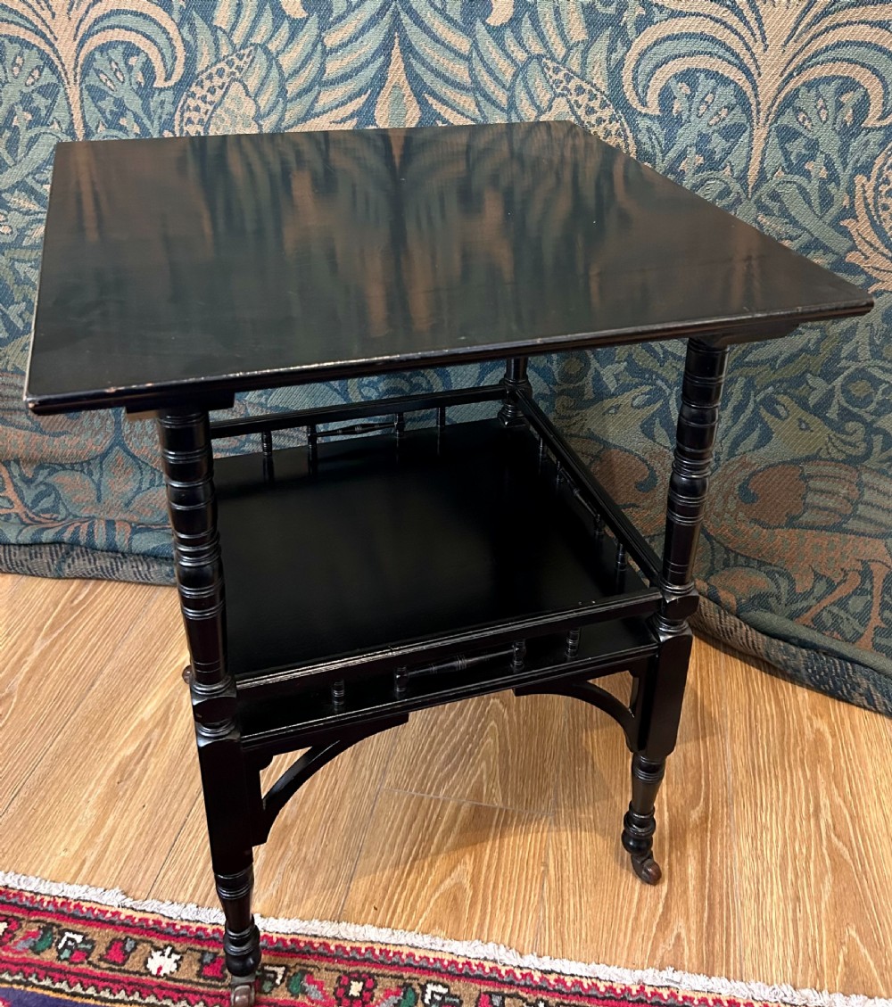 An Aesthetic Movement ebonised wooden side table featuring turned legs, a lower shelf with spindle railing, arched corner brackets, and brass castors. View IV