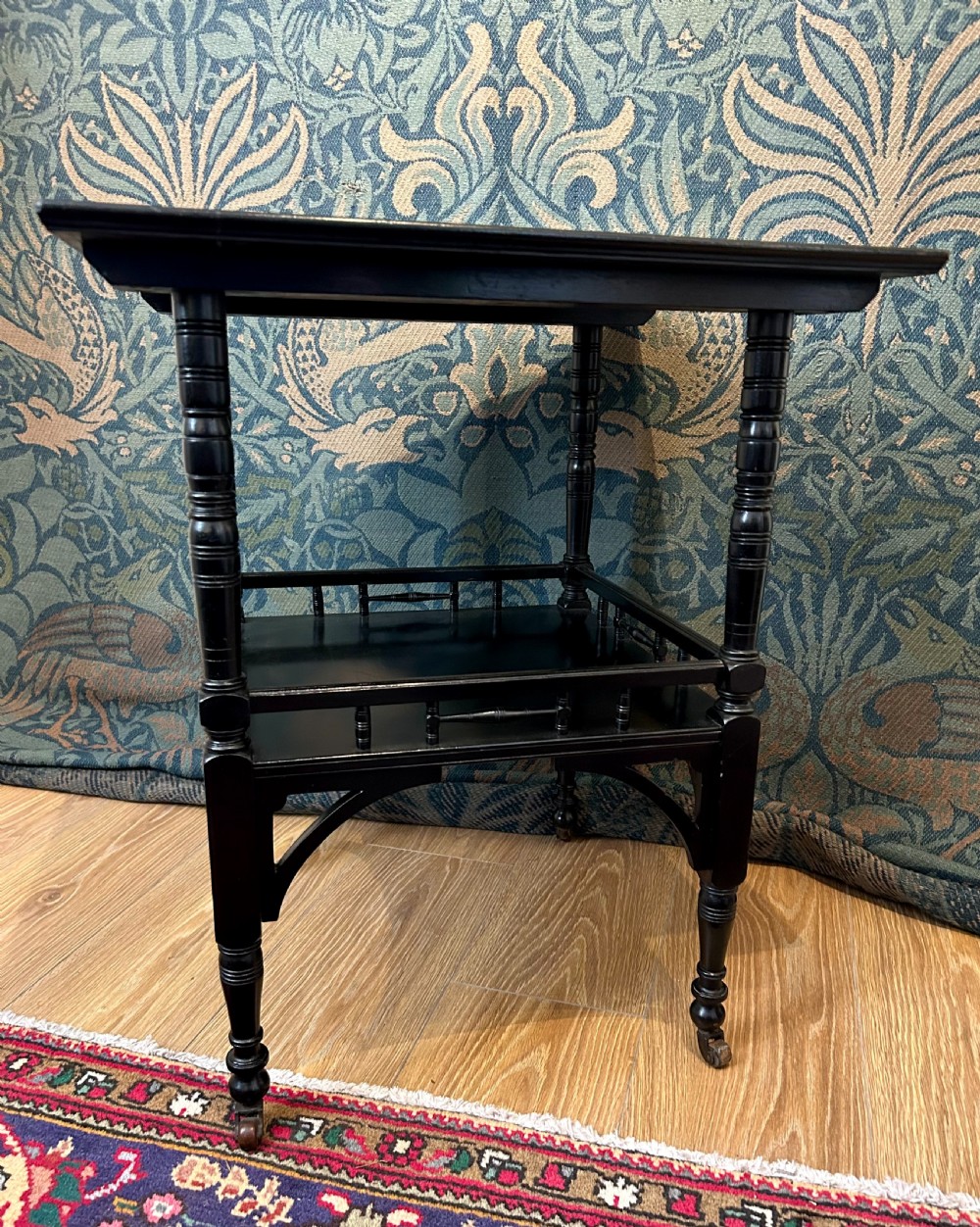 An Aesthetic Movement ebonised wooden side table featuring turned legs, a lower shelf with spindle railing, arched corner brackets, and brass castors. View V