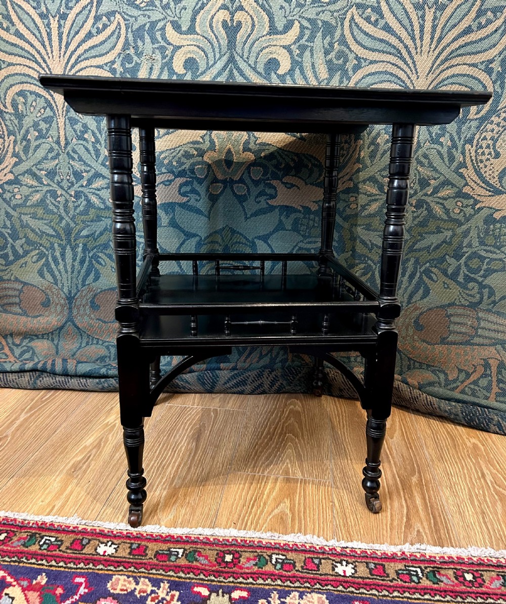 An Aesthetic Movement ebonised wooden side table featuring turned legs, a lower shelf with spindle railing, arched corner brackets, and brass castors. View VI