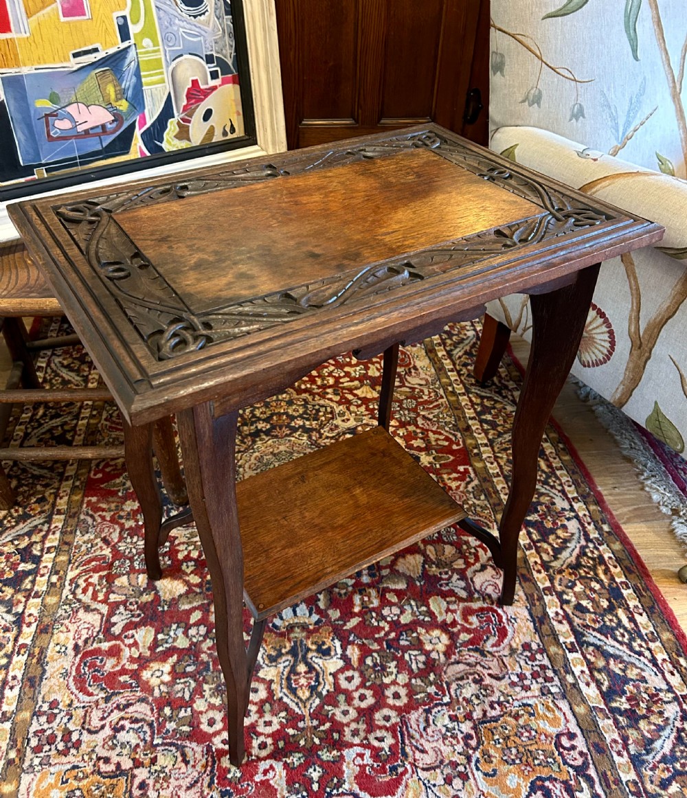 A warm brown oak occasional table with a relief-carved botanical border on the tabletop, slender curved legs, and an integrated lower rectangular tier.