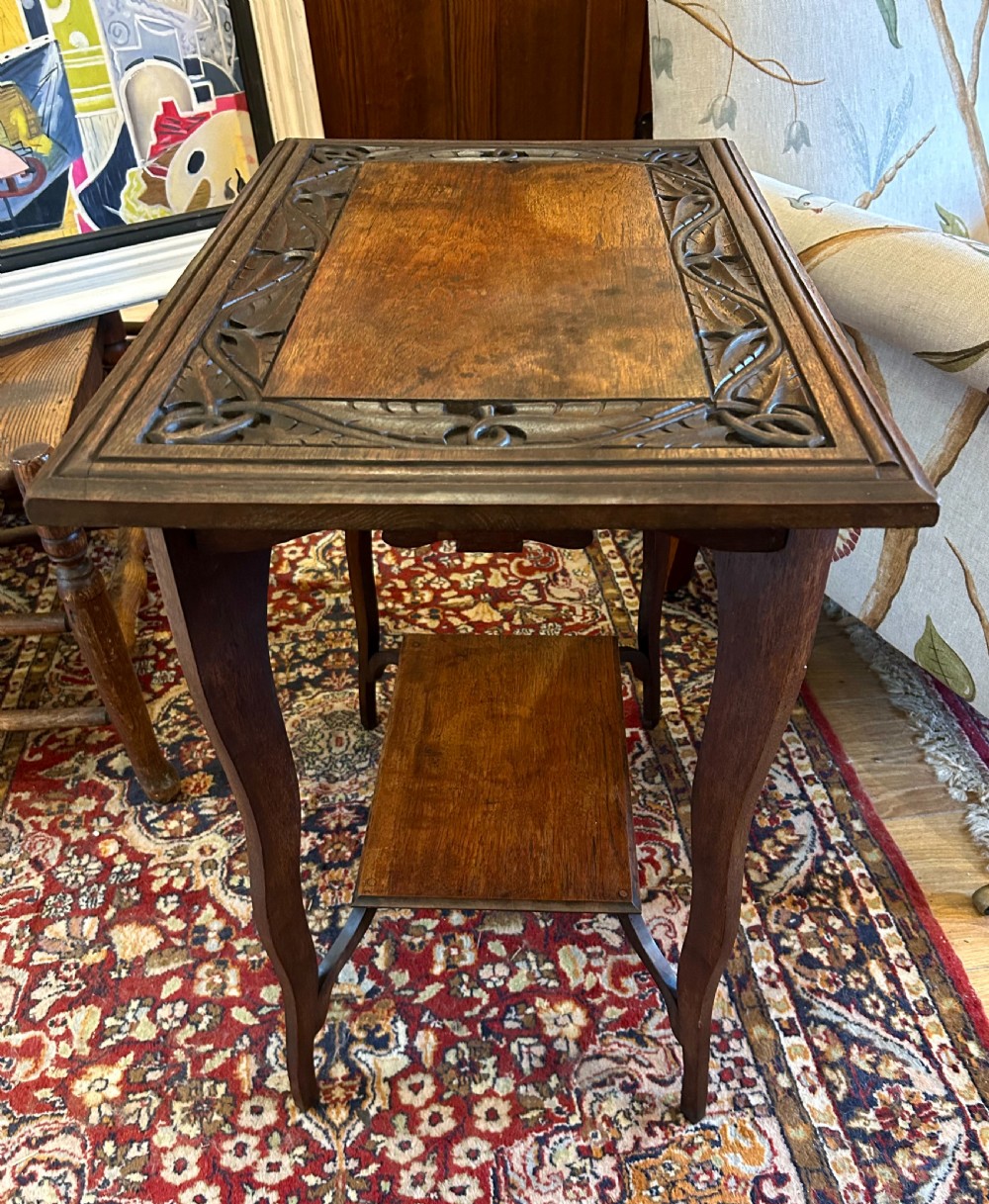 A warm brown oak occasional table with a relief-carved botanical border on the tabletop, slender curved legs, and an integrated lower rectangular tier. View II