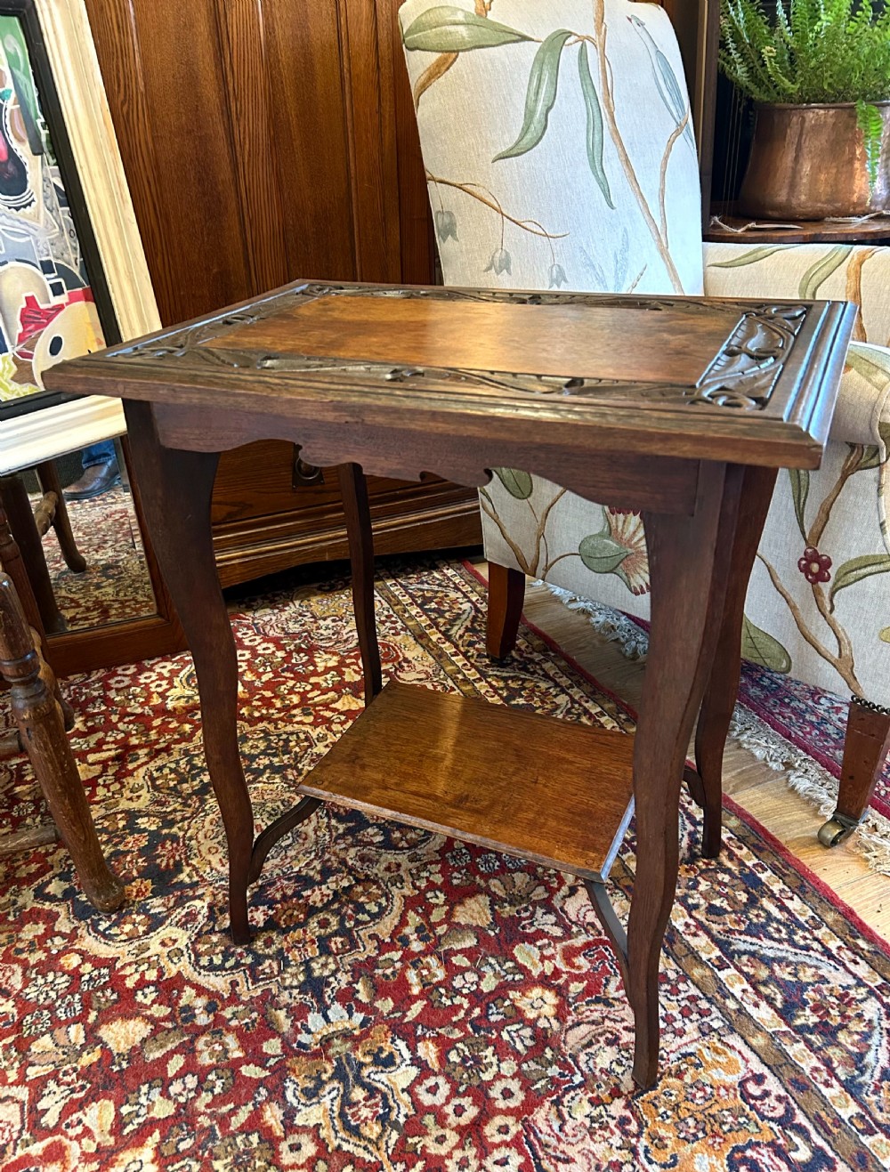 A warm brown oak occasional table with a relief-carved botanical border on the tabletop, slender curved legs, and an integrated lower rectangular tier. View III