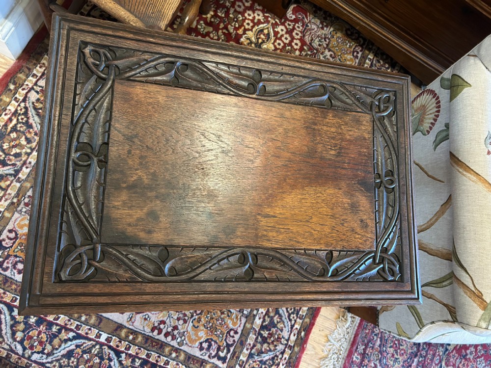 A warm brown oak occasional table with a relief-carved botanical border on the tabletop, slender curved legs, and an integrated lower rectangular tier. View VI