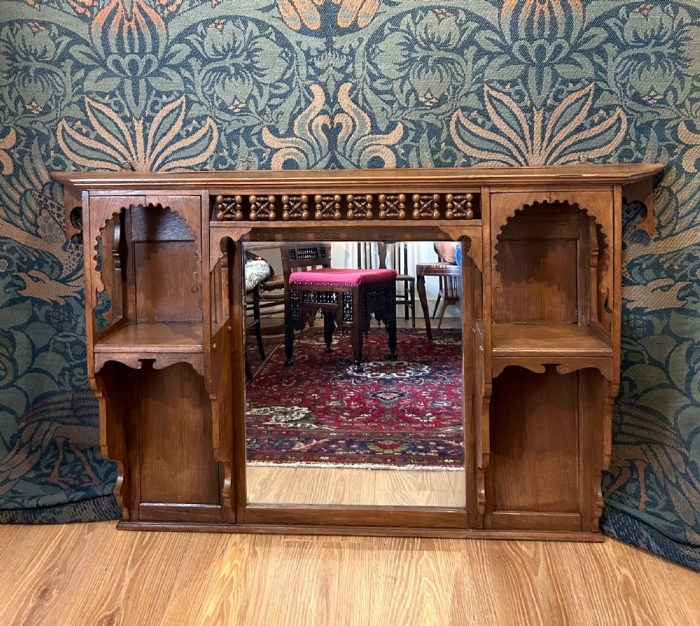 A carved wooden Moorish style overmantel mirror with a central glass panel, horseshoe arch side shelves, and a top turned spindle gallery.