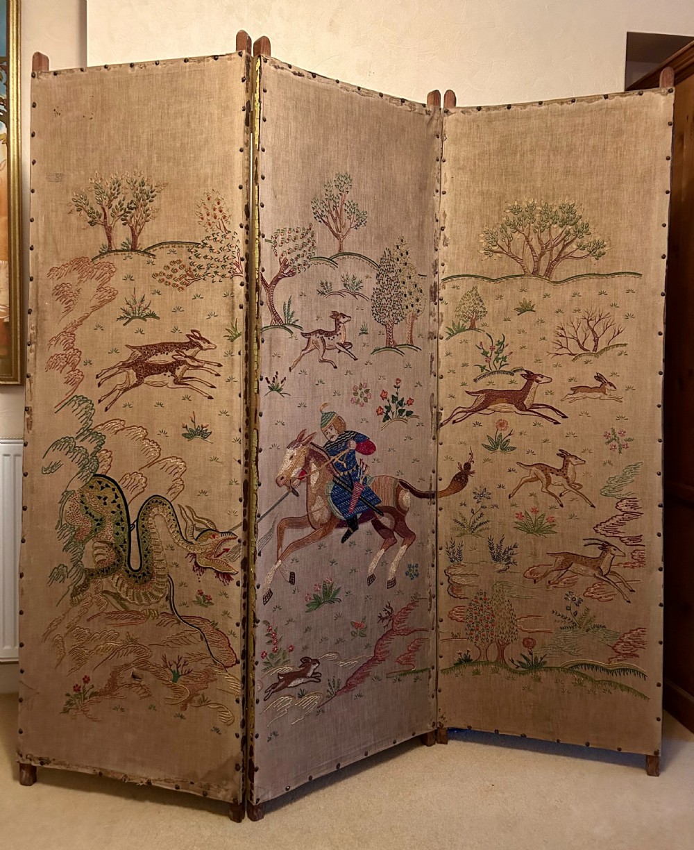 A three-panel folding room screen constructed with natural beige linen panels embroidered with Persian-style equestrian and mythological creature motifs, secured with brass tacks.