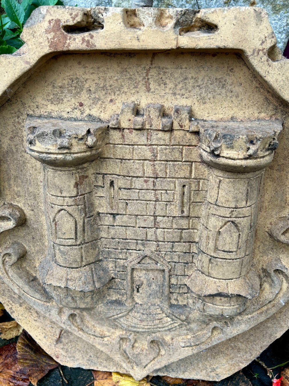 A carved buff stone armorial shield plaque featuring a relief depiction of a twin-towered castle gate with scrolled side details. View IV
