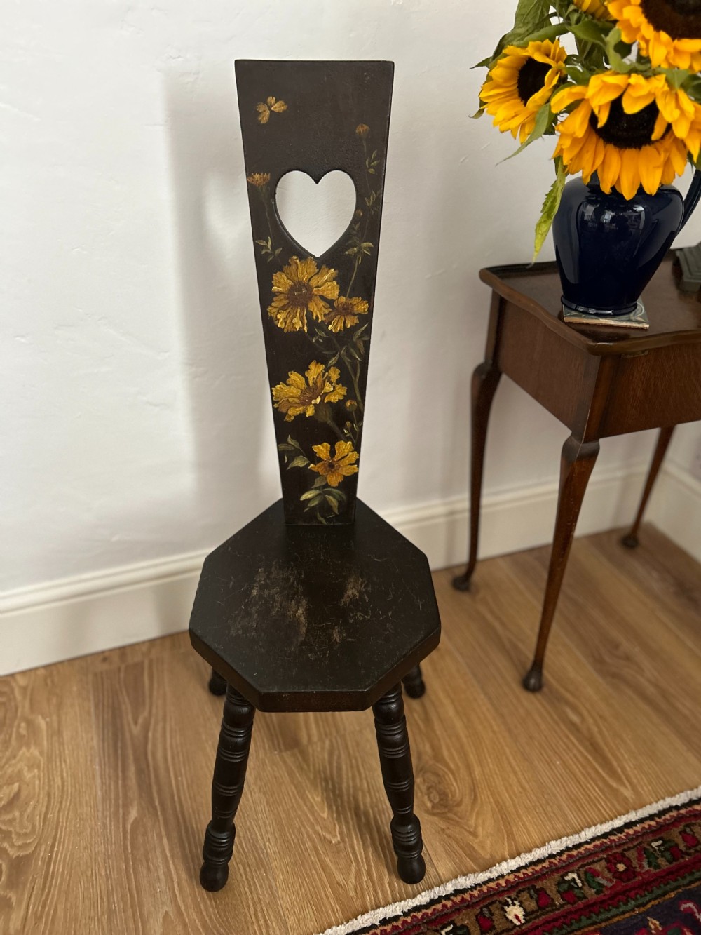 A dark wood Welsh spinning chair featuring a tall narrow backrest decorated with hand-painted yellow flowers and a heart cutout above a hexagonal seat and turned legs.