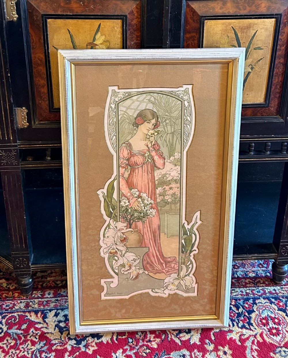 A framed Art Nouveau lithograph depicting a woman in a long pink dress holding flowers inside a conservatory setting with a botanical border.