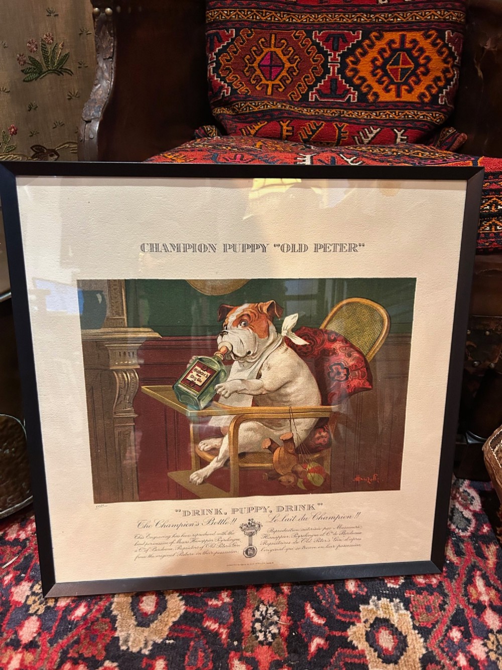 A framed 1920s French advertising print titled Drink, Puppy, Drink depicting a bulldog seated in a high chair drinking from a bottle.