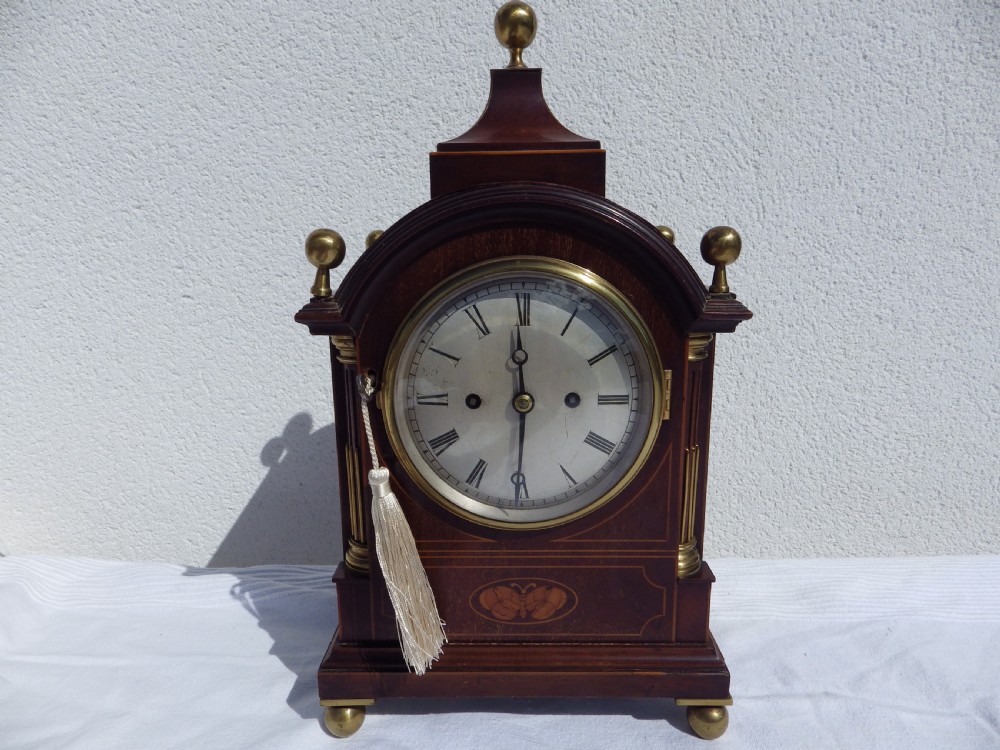 An English double fusee bracket clock crafted from dark mahogany wood with brass finials, brass column accents, marquetry inlay, and a circular white dial featuring Roman numerals.