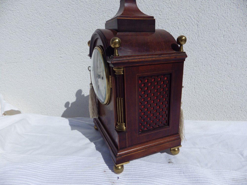 An English double fusee bracket clock crafted from dark mahogany wood with brass finials, brass column accents, marquetry inlay, and a circular white dial featuring Roman numerals. View IV