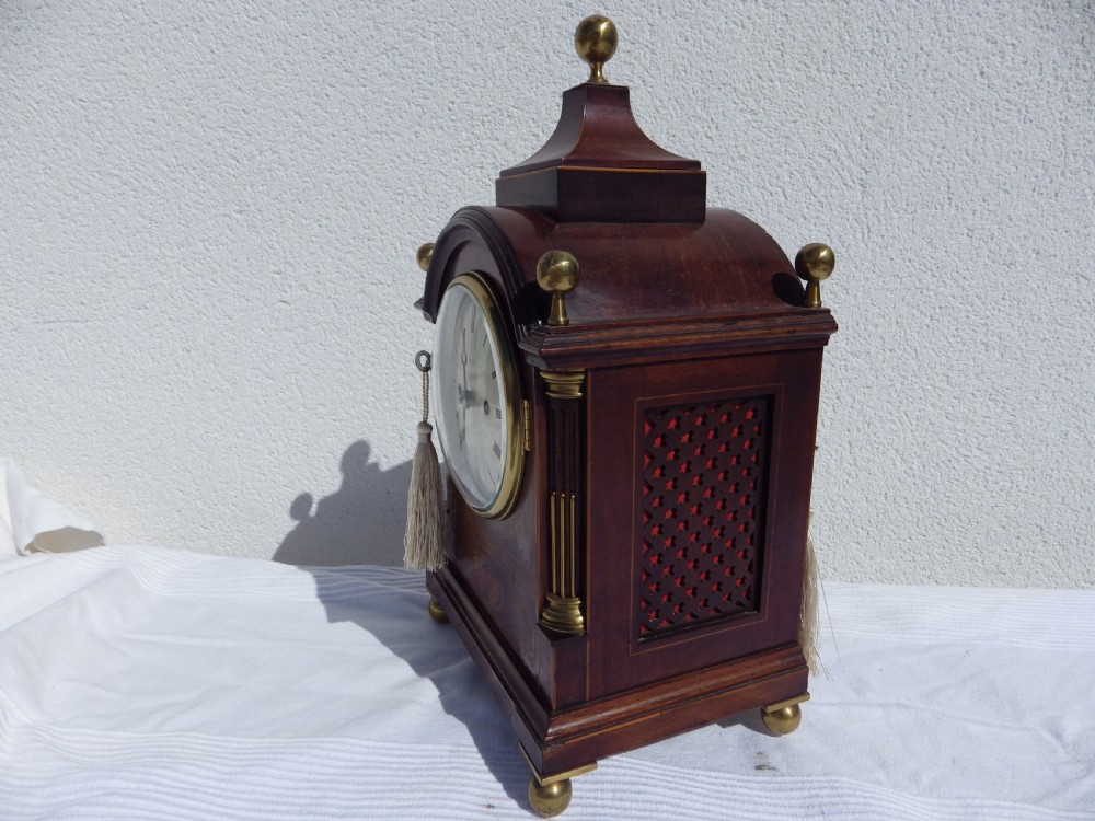 An English double fusee bracket clock crafted from dark mahogany wood with brass finials, brass column accents, marquetry inlay, and a circular white dial featuring Roman numerals. View V