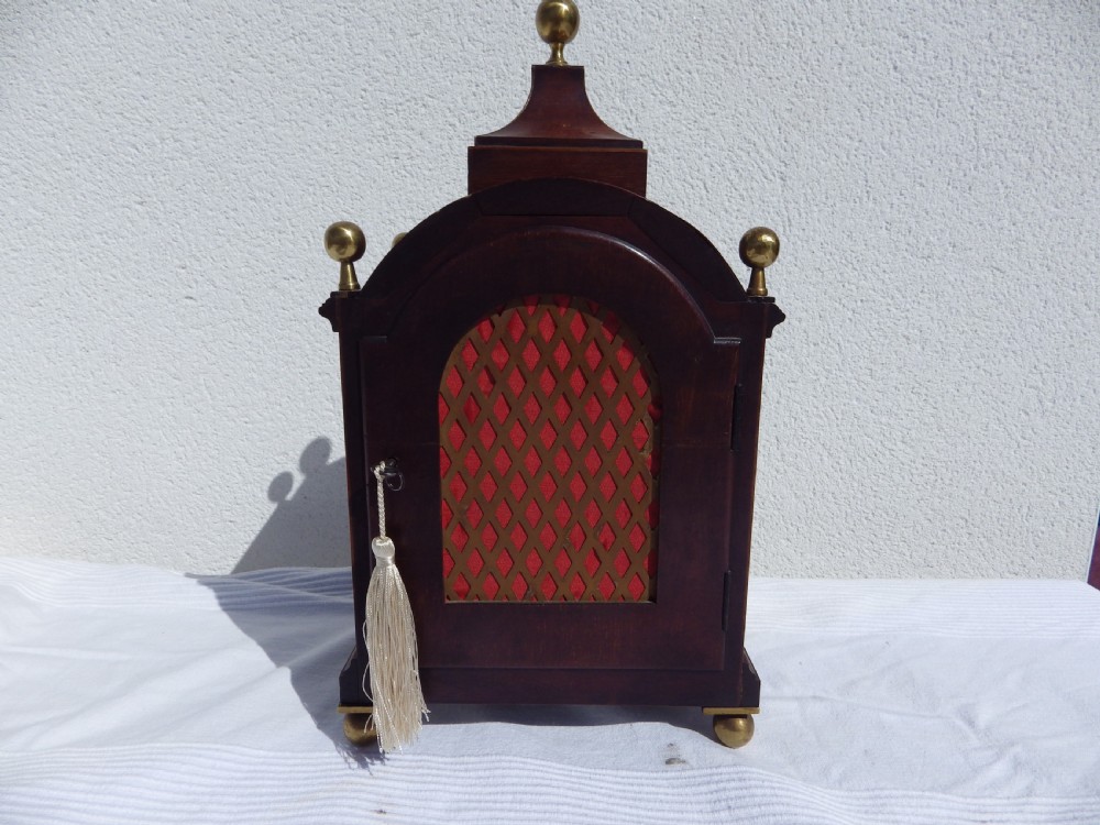 An English double fusee bracket clock crafted from dark mahogany wood with brass finials, brass column accents, marquetry inlay, and a circular white dial featuring Roman numerals. View VI