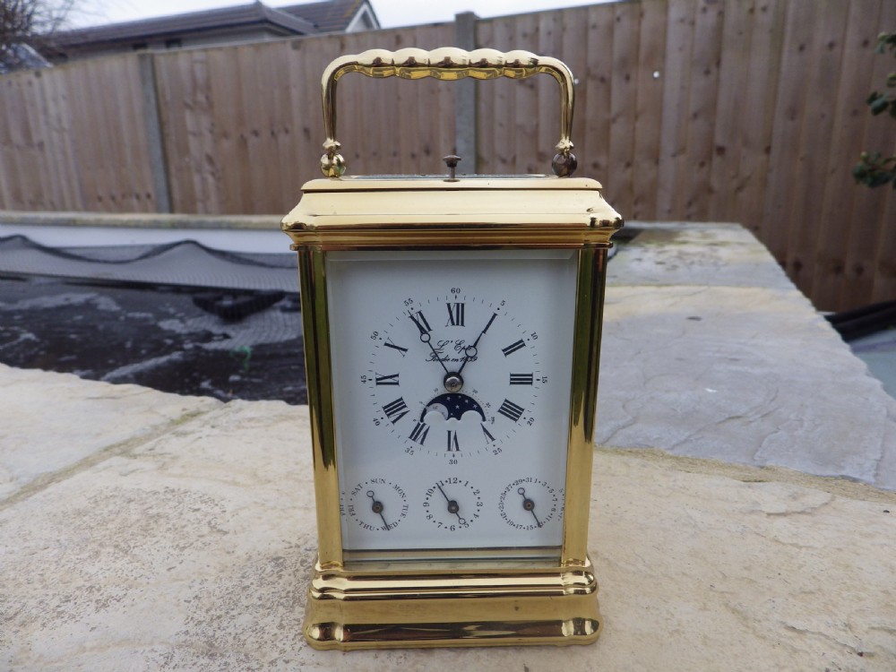 A polished brass LEpee carriage clock featuring bevelled glass panels, a white dial with Roman numerals, a moon phase aperture, and three subsidiary calendar dials.
